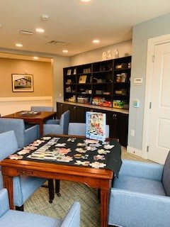 A cozy library space featuring a puzzle in progress on a wooden table surrounded by comfortable seating and bookshelves filled with various items.