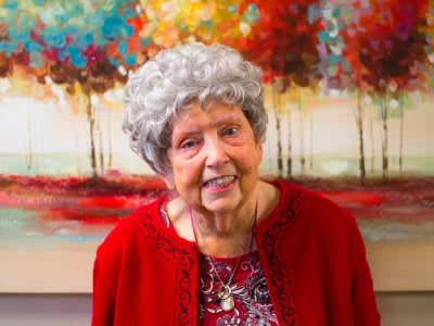 An elderly woman smiling warmly in front of a colorful painting, wearing a red outfit.