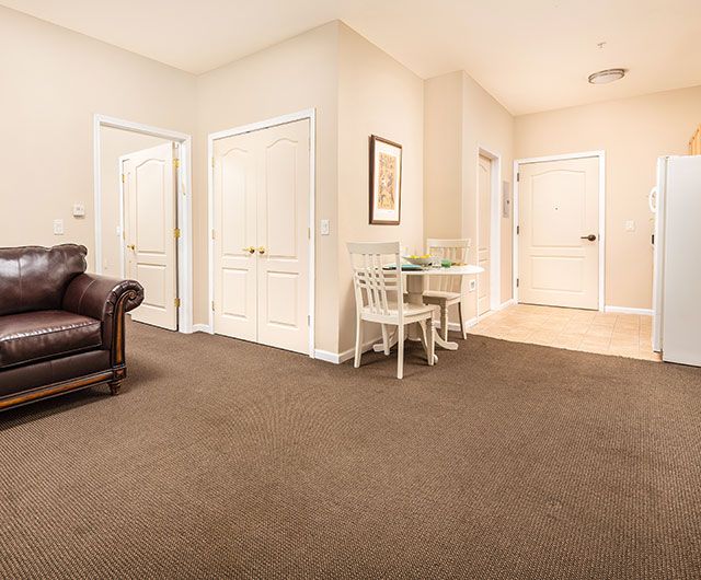 A cozy living room with beige walls, a brown carpet, and white doors. The space includes a leather sofa and a small table with two chairs.