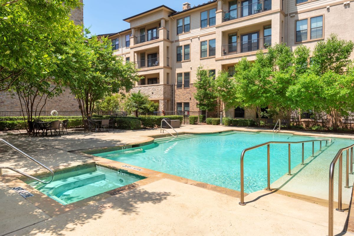 An outdoor swimming pool adjacent to an apartment building, surrounded by trees and landscaped areas.