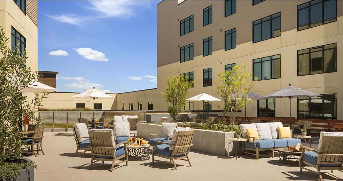 A well-furnished outdoor patio area with comfortable seating and umbrellas, adjacent to a multi-story building.