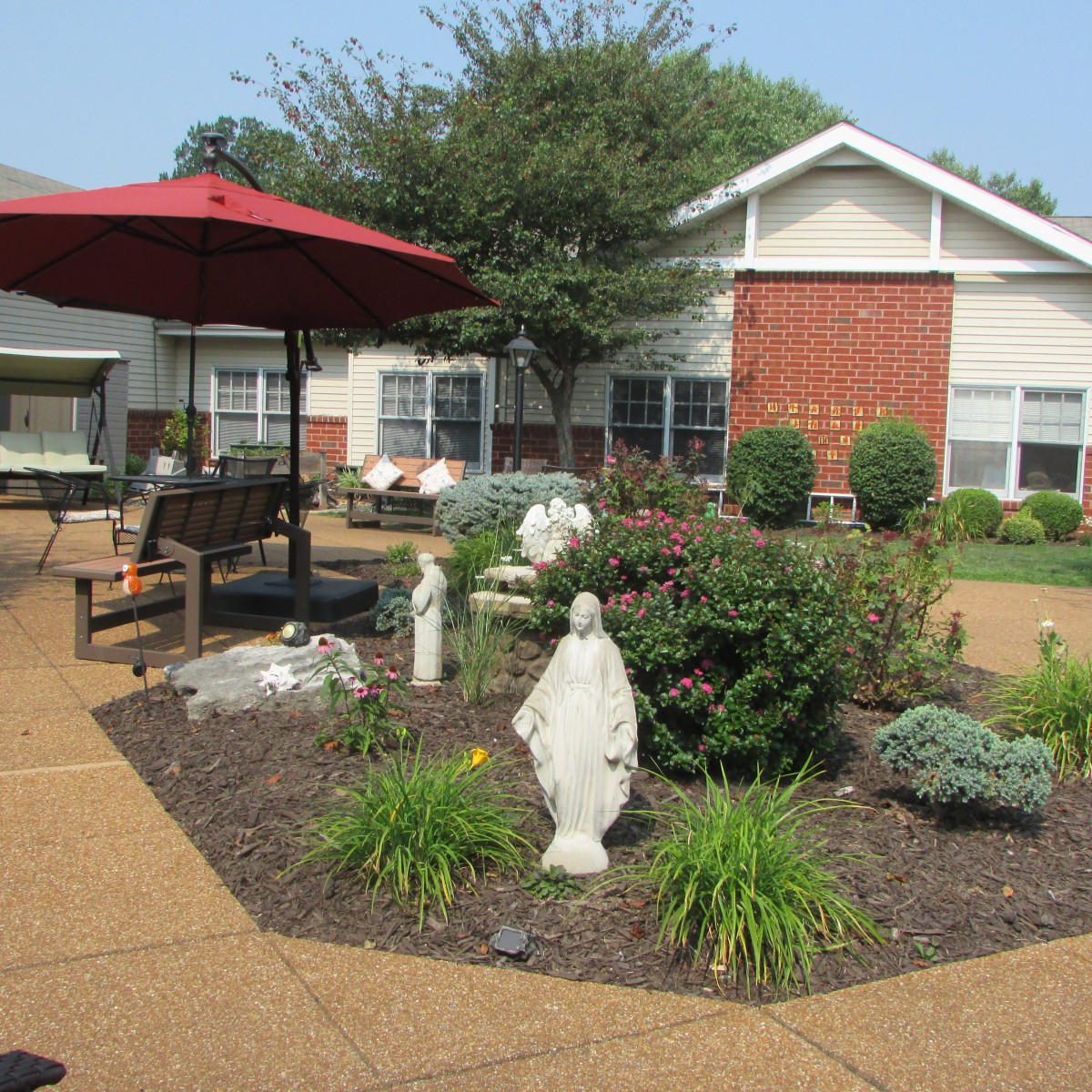 A well-maintained garden with a central statue, surrounded by benches and an umbrella on a patio, providing a peaceful outdoor setting.