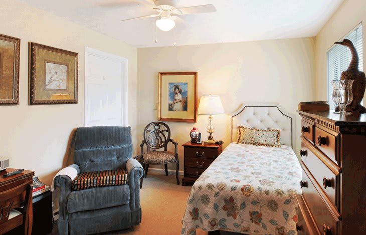 A cozy bedroom featuring a single bed, an armchair, and a nightstand with a lamp. The walls are adorned with decorative artworks.