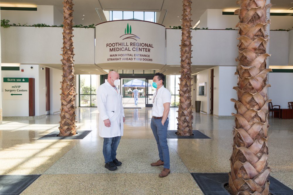 Two individuals, one in a lab coat, converse in the lobby of a medical center with palm trees and an open entrance.
