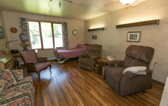 A cozy nursing home room with a bed, recliner, and large window.