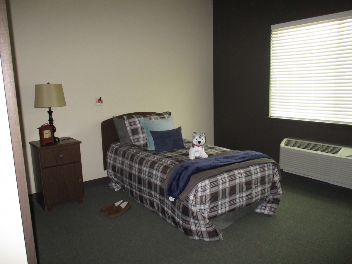 A simple bedroom with a single bed covered in plaid bedding, accompanied by a nightstand and lamp.