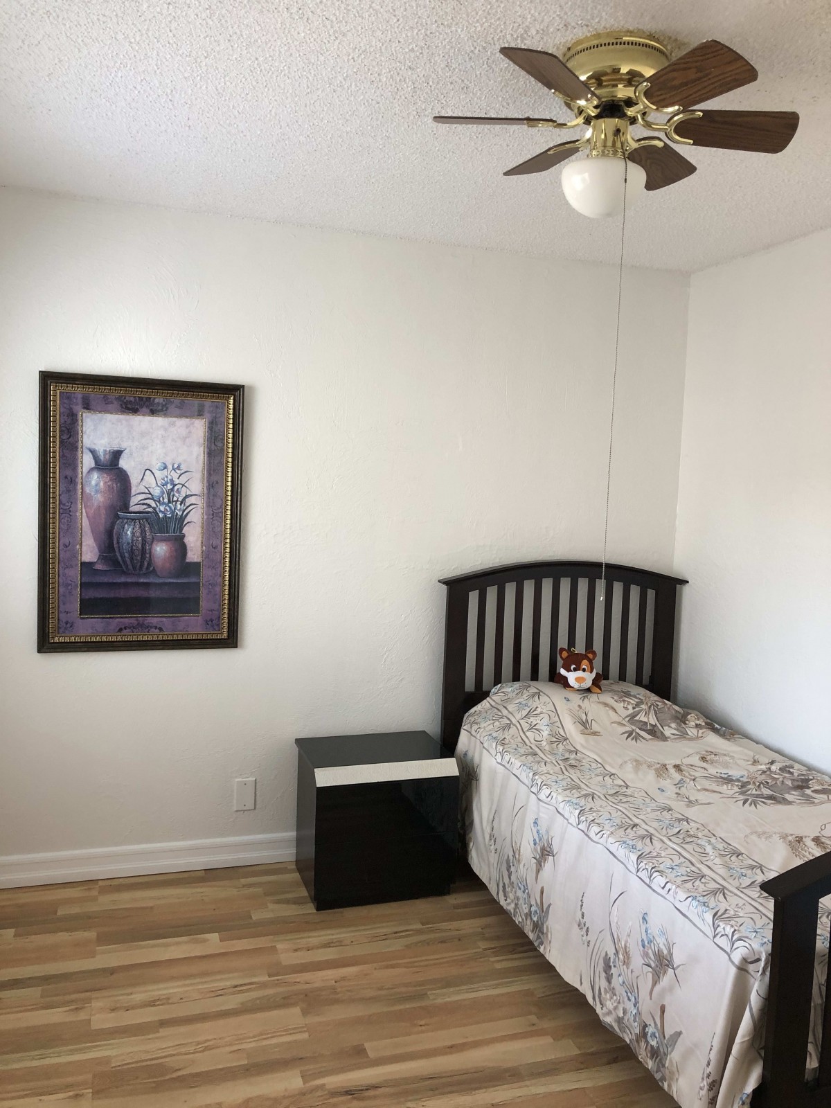 A minimalist bedroom featuring a single bed, nightstand, ceiling fan, and framed wall art.