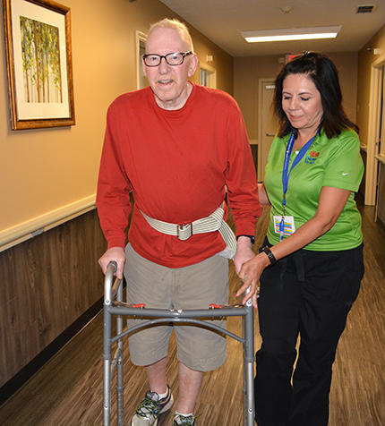An elderly man using a walker is assisted by a caregiver in a hallway.