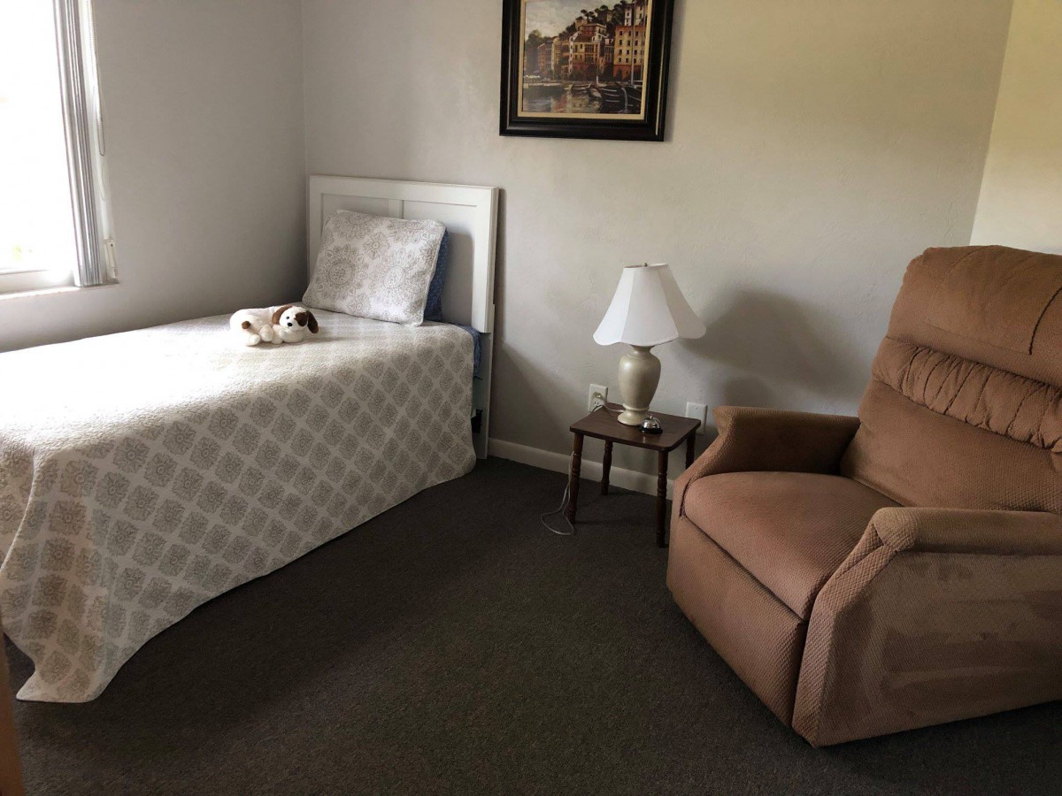 A simple bedroom with a single bed, a lamp on a side table, and an upholstered chair.