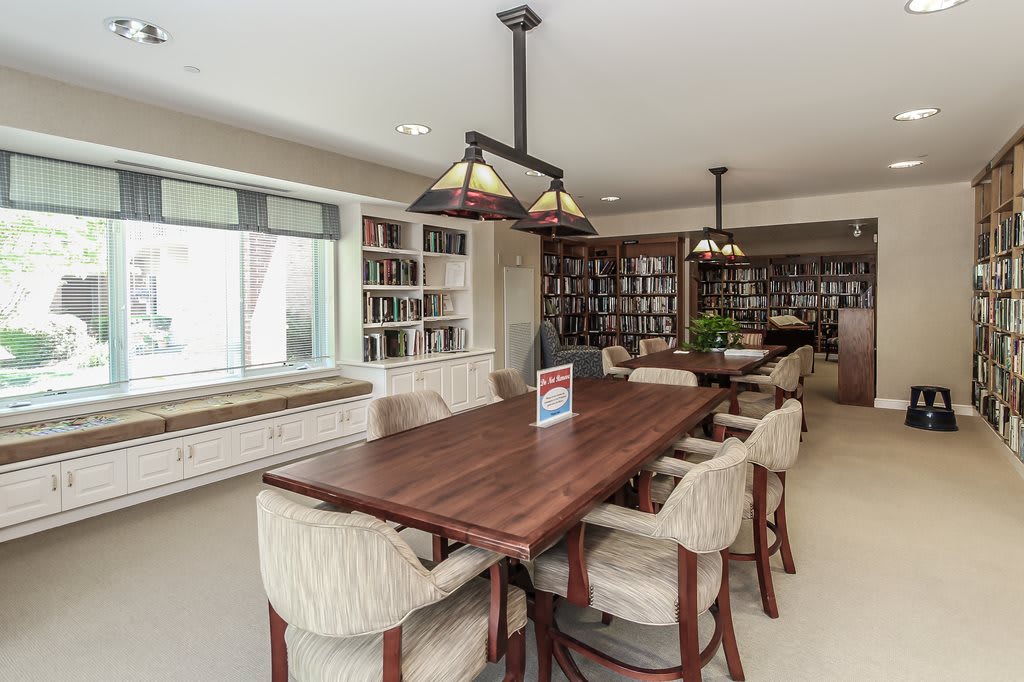 A cozy library with a large wooden dining table surrounded by plush chairs and bookshelves filled with books.