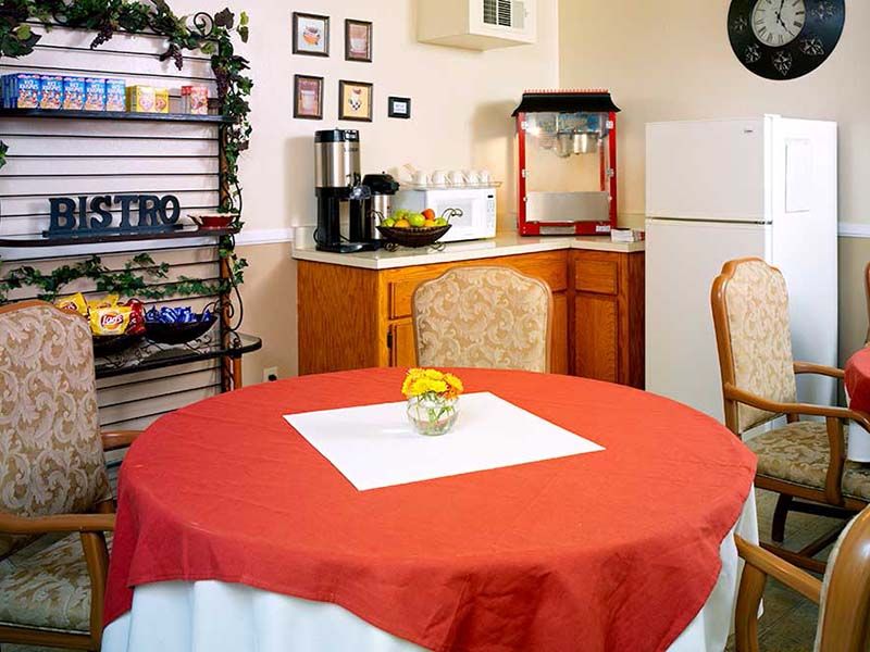 A cozy kitchen setup featuring a table with a red tablecloth, chairs, and various appliances including a microwave, popcorn machine, and coffee maker.