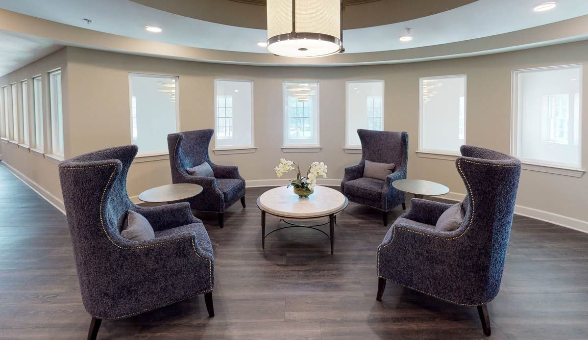 A modern lounge area with comfortable armchairs arranged around a central coffee table, illuminated by natural light from multiple windows.