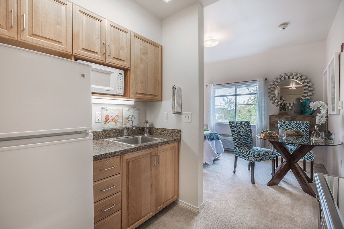 A compact kitchen in an apartment features wooden cabinets, a microwave, and a refrigerator opening into a cozy dining area with large windows.