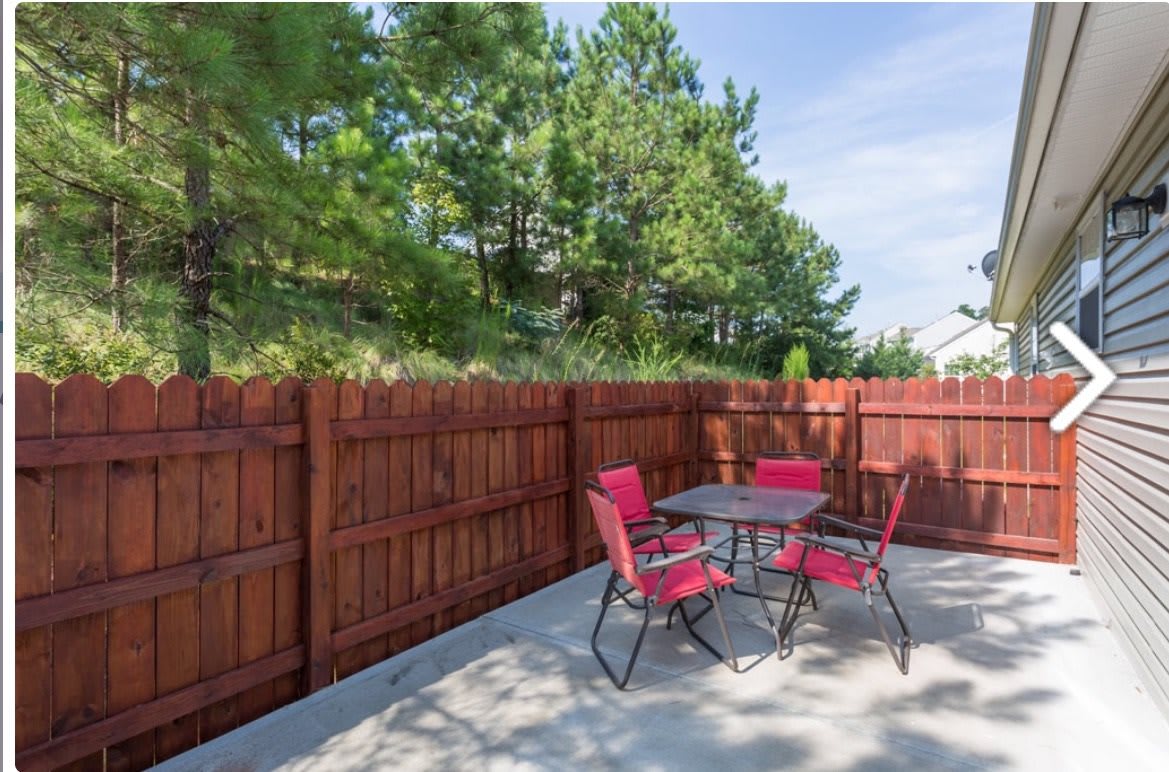 A sunny backyard patio with red chairs and a table next to a wooden fence.