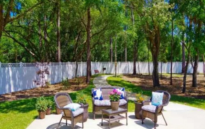A tranquil backyard patio with seating surrounded by lush greenery and a white fence.
