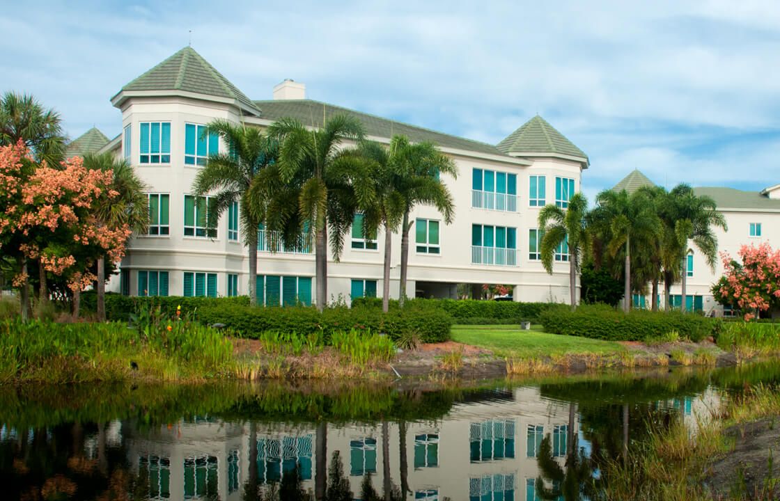 A modern building surrounded by palm trees, reflected in a calm lake.
