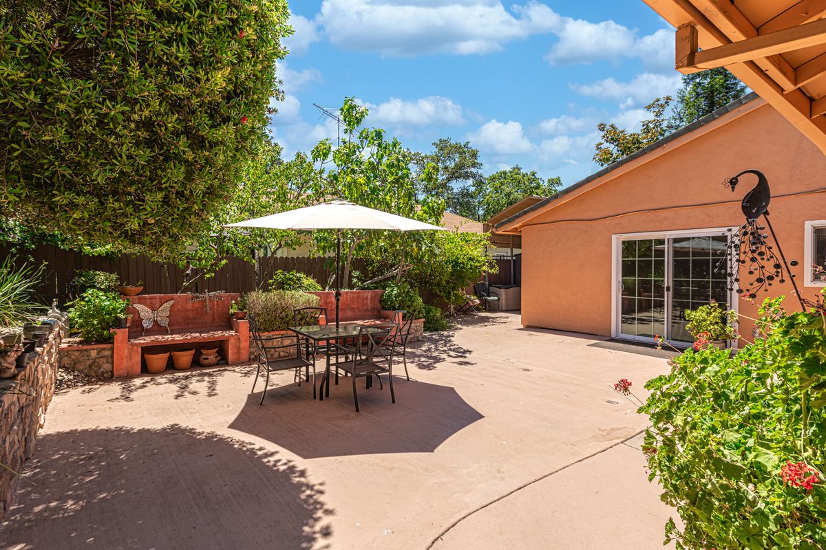 A spacious backyard patio featuring outdoor seating under a large umbrella, surrounded by lush greenery.