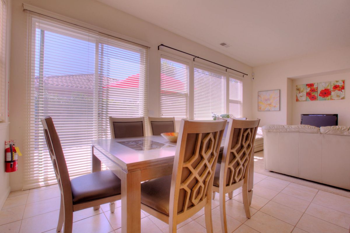 A bright dining room with a wooden table and chairs, large windows, and blinds.