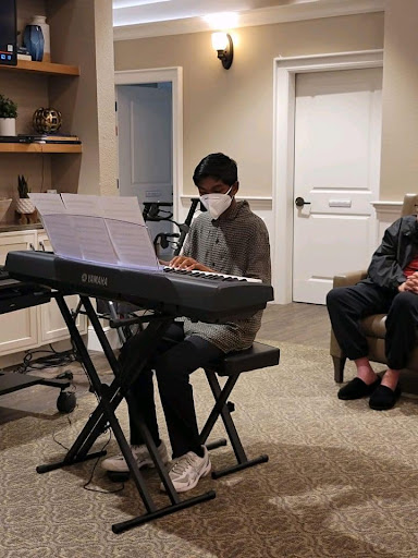 A young musician performs on a digital piano while wearing a mask, with an audience member seated nearby.