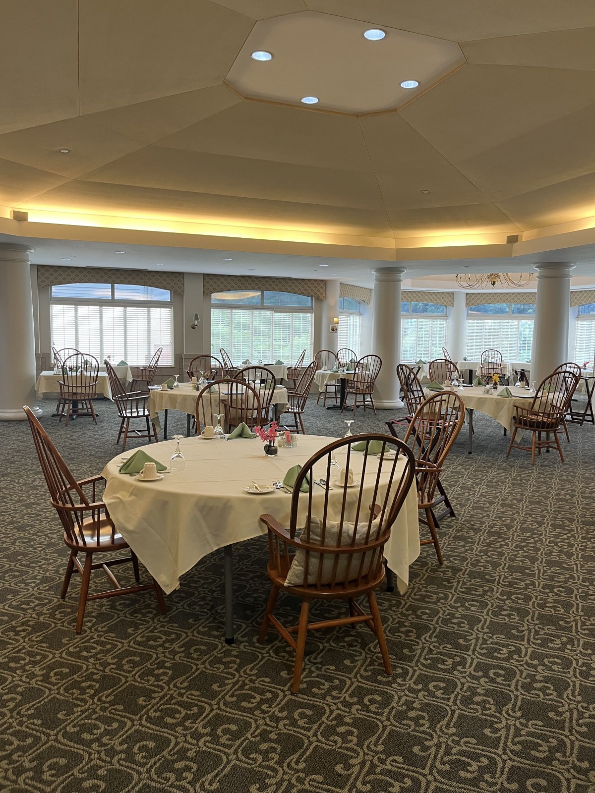 An empty dining room with round tables and chairs, featuring large windows and a carpeted floor.