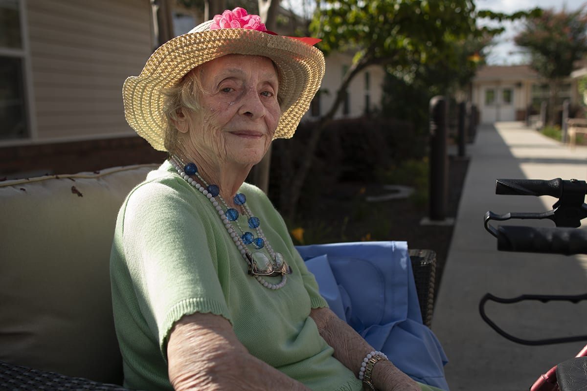 An elderly woman wearing a straw hat sits outdoors in the sunlight, appearing relaxed and content.