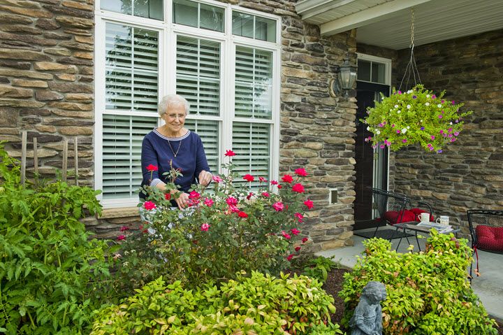 An elderly woman tending to her garden in front of a brick house.