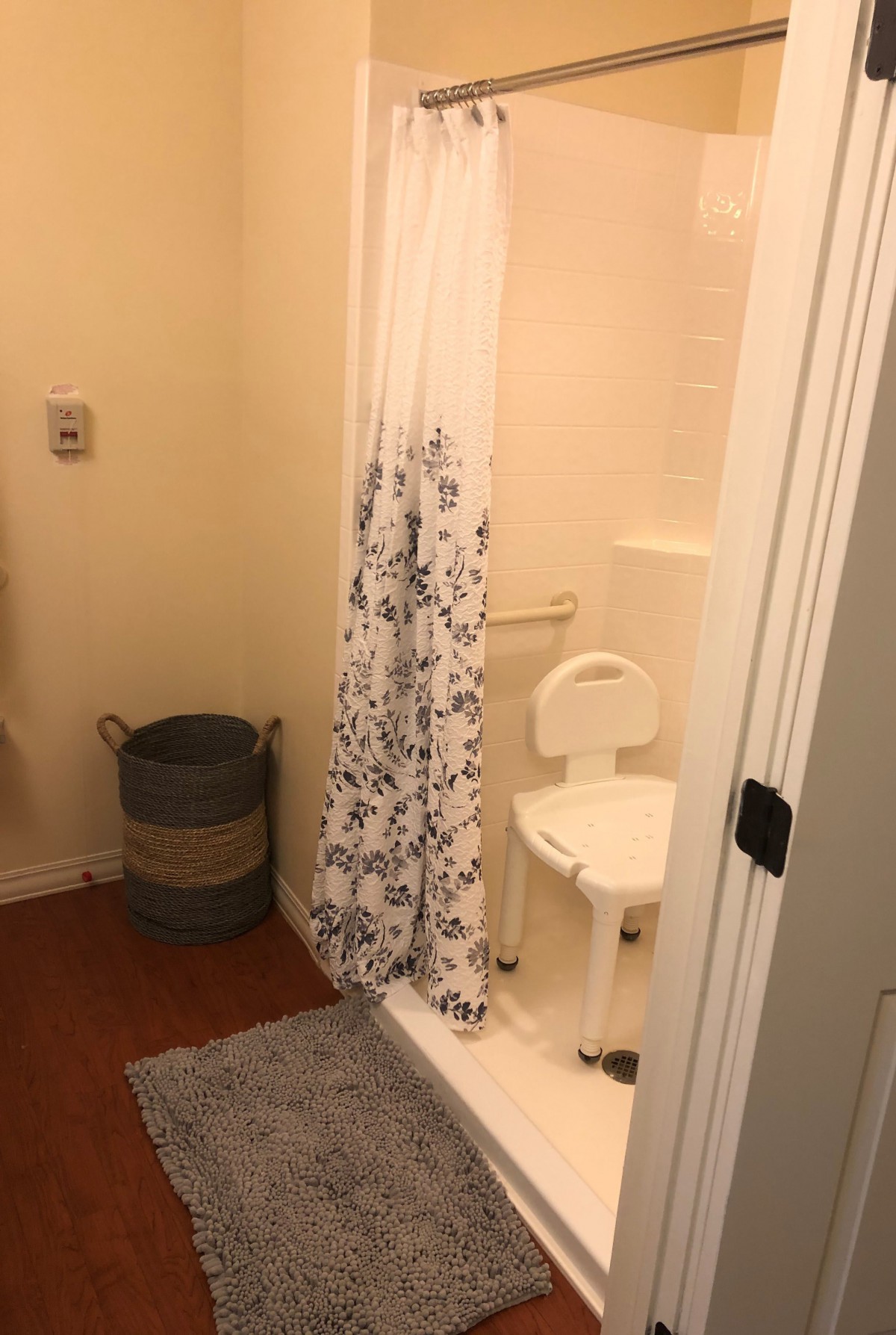 A bathroom with a shower featuring a floral curtain, a shower chair, and a laundry basket.