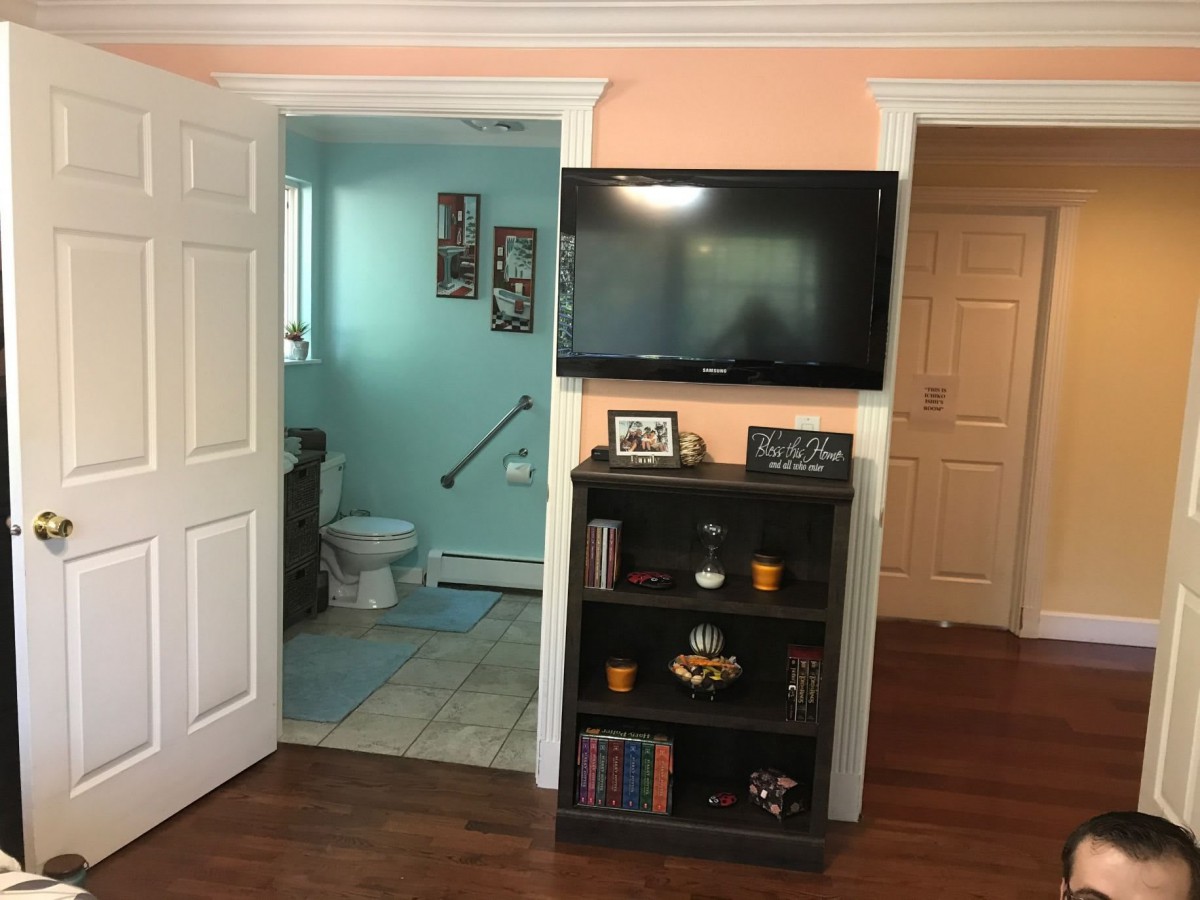 A cozy living space featuring a wall-mounted television, a bookshelf stocked with books and decorative items, and a glimpse into an adjacent bathroom.