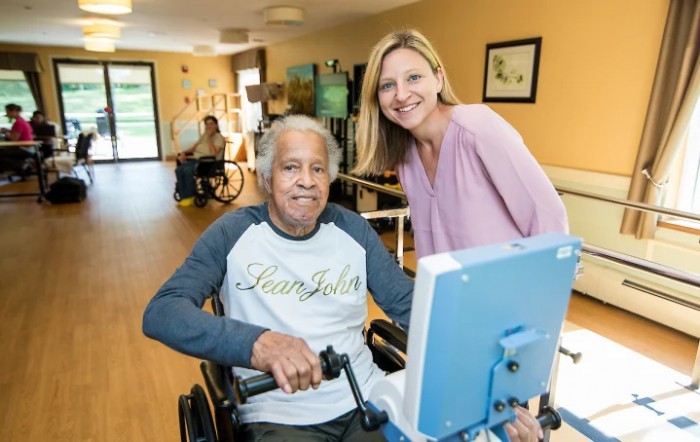 An elderly man in a wheelchair using exercise equipment with assistance from a healthcare worker in a nursing home.
