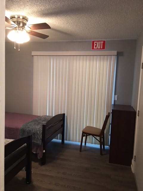 A small bedroom with a bed, dresser, chair, and sliding door with vertical blinds under a ceiling fan.