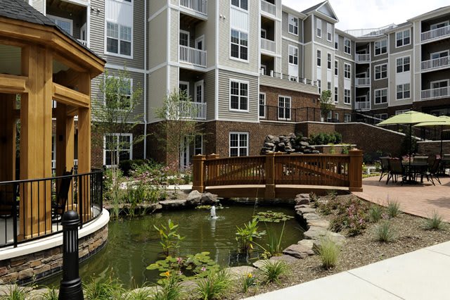 A modern apartment complex with a landscaped pond and wooden bridge in the courtyard.