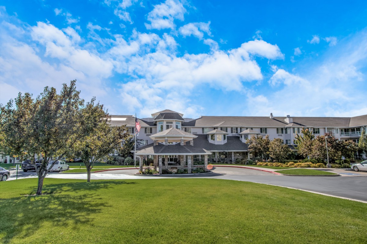 A large residential building surrounded by a well-maintained lawn and trees under a bright blue sky.