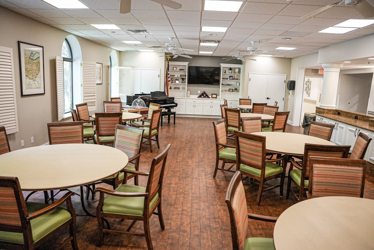 A well-lit dining room featuring round tables and chairs, a television, and a piano.