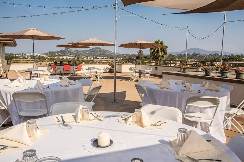 A rooftop dining area with tables set under umbrellas on a sunny day, overlooking a scenic landscape.