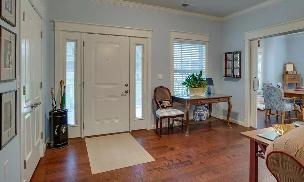 A welcoming home entrance with a hardwood floor, featuring a front door flanked by two narrow windows. A decorative table with a plant and lamp adds charm, accompanied by an elegant chair.