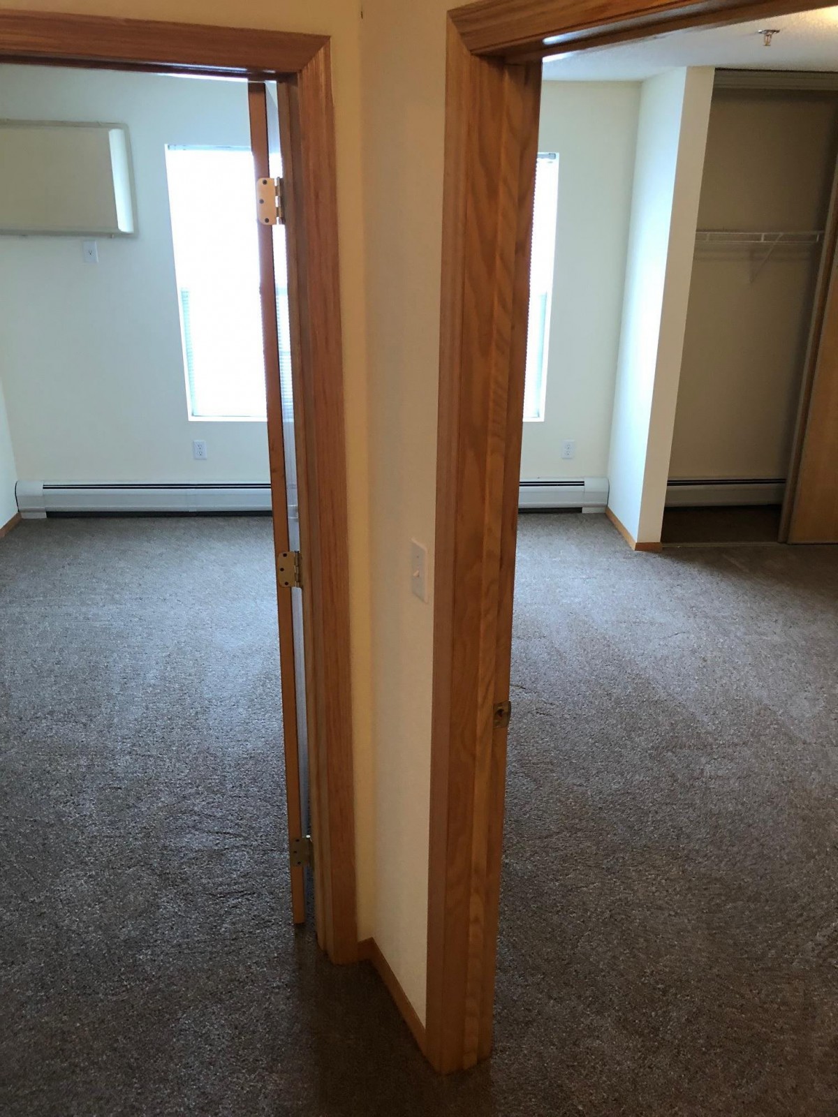 An interior view of an empty room with a door frame separating two carpeted areas, illuminated by natural light from the windows.