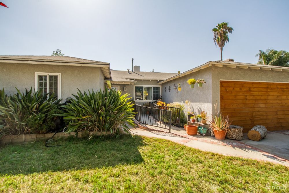 A suburban house with a landscaped yard and a pathway leading to the entrance, featuring various plants and potted decorations under a clear sky.