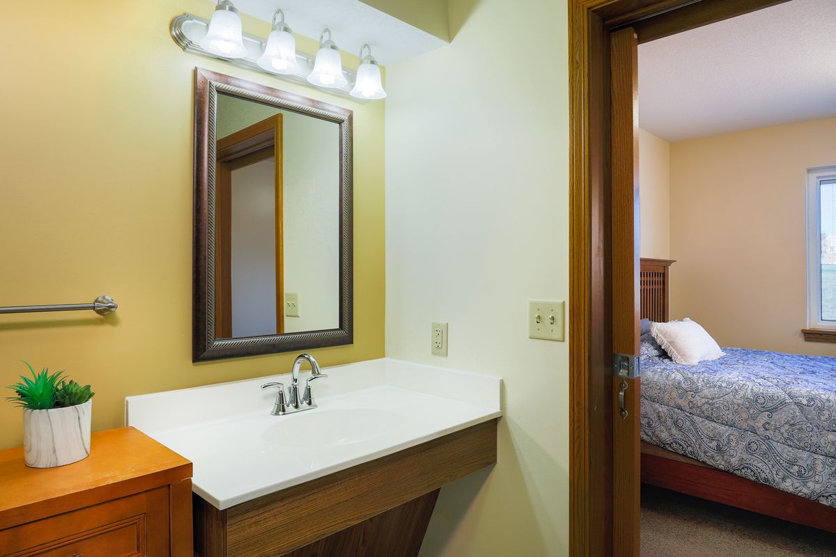 A cozy bathroom with a mirror, sink, and overhead lighting adjacent to a bedroom.
