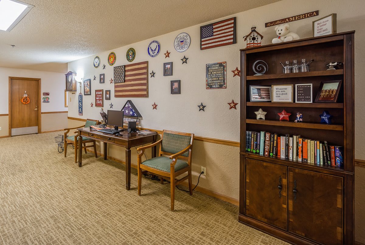 A cozy office space featuring a desk with a computer and chairs, complemented by a bookshelf filled with books and decorative items. The walls are adorned with American flags and various plaques.
