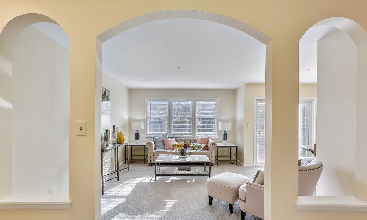 A bright and elegantly furnished living room viewed through an archway, with large windows allowing natural light to fill the space.