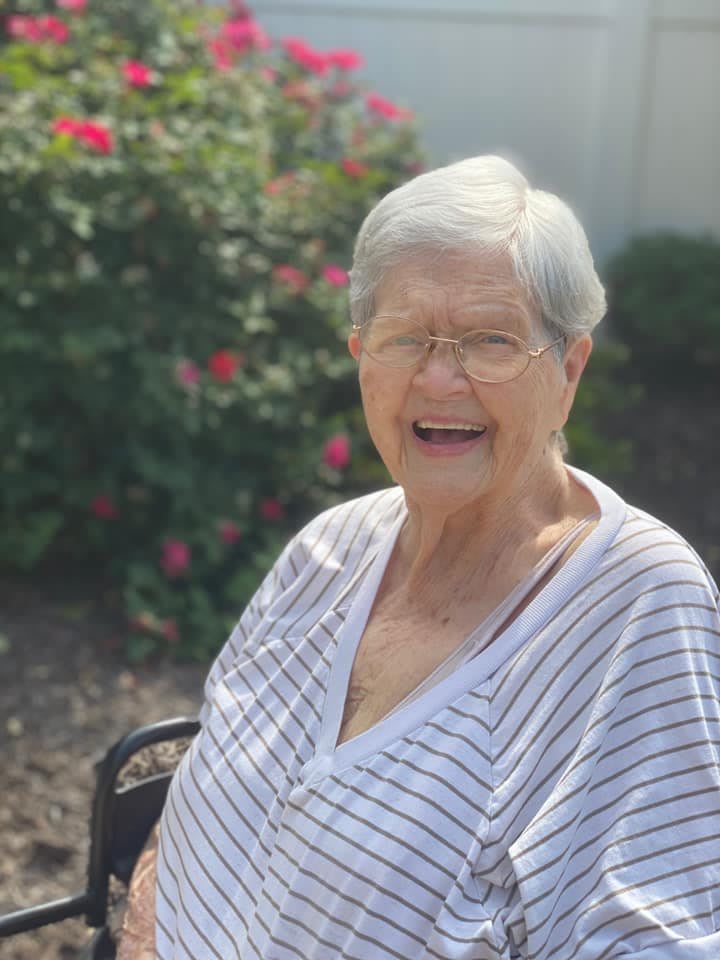 An elderly woman smiling while sitting outdoors in a garden setting.