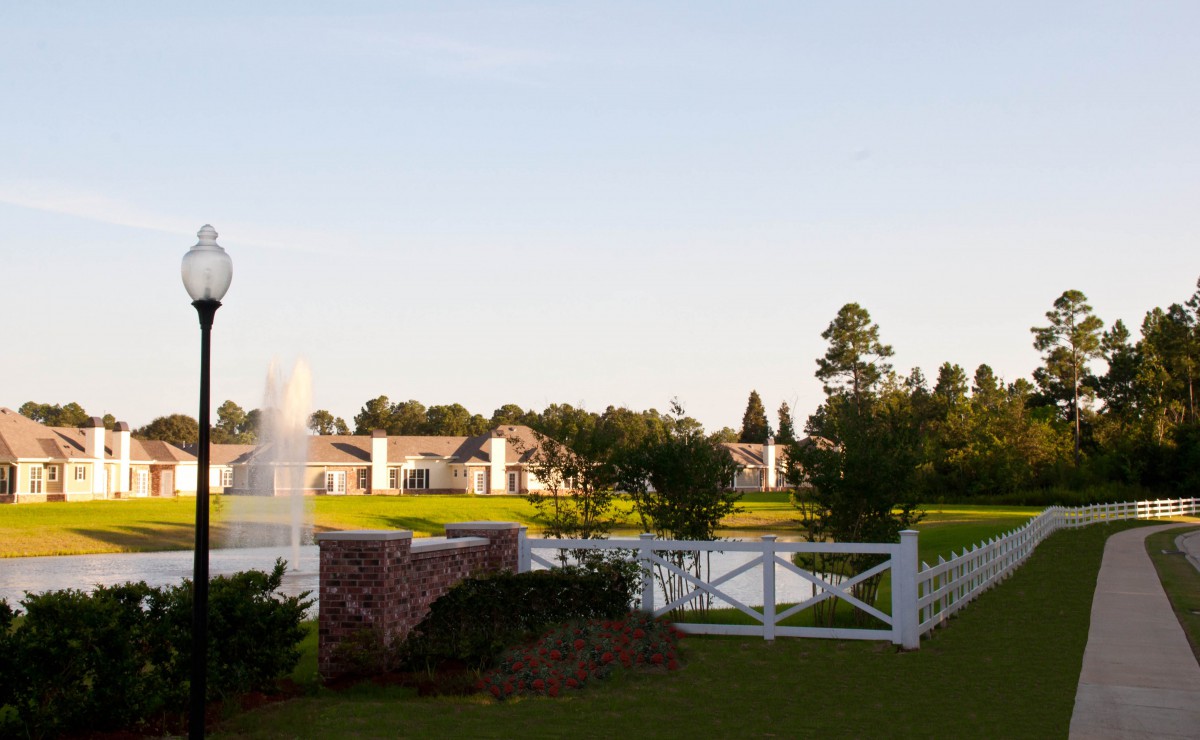 A tranquil landscape featuring a small lake with a fountain, surrounded by houses and trees, with a white fence in the foreground.