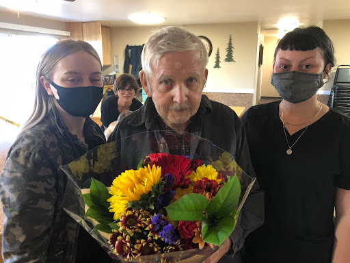 An elderly man surrounded by two individuals, holding a vibrant bouquet of flowers in a warm indoor setting.