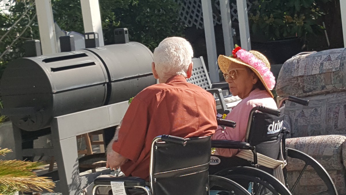 An elderly couple in wheelchairs enjoying a sunny day outdoors near a grill.