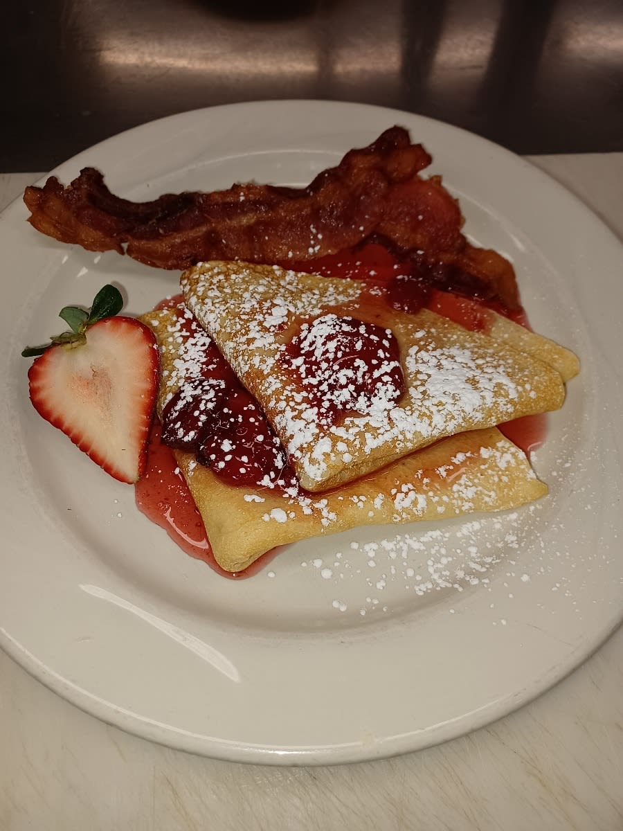 A delicious breakfast plate featuring crepes topped with strawberries and powdered sugar, served with crispy bacon.