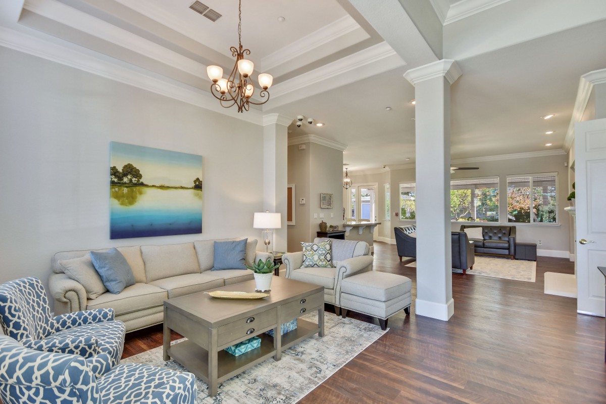 A spacious living room featuring elegant interior design with a large sofa, coffee table, and chandelier, highlighted by artwork on the wall and hardwood flooring.