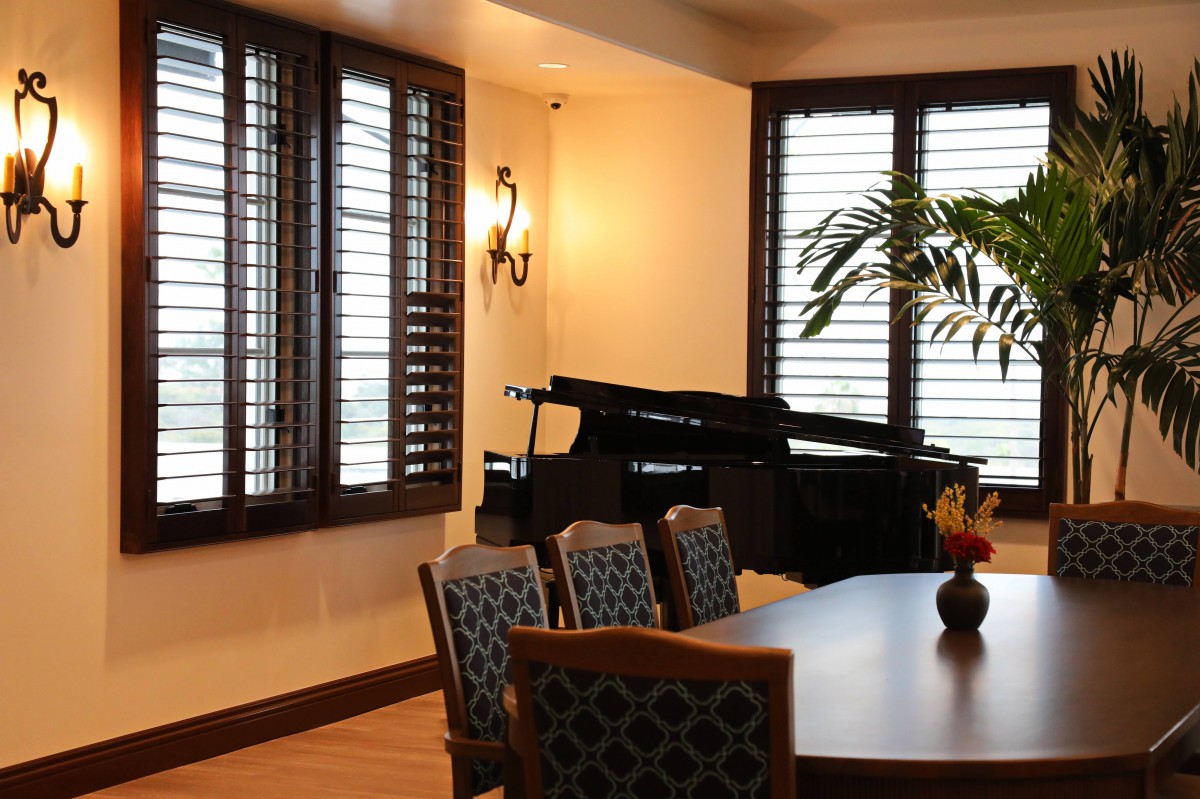 A cozy dining room with a grand piano and elegant furnishings near large windows with shutters.
