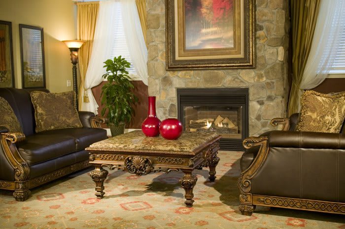 A cozy living room featuring an ornate coffee table, framed by sofas and a stone fireplace, with decorative elements including red vases and a large painting.