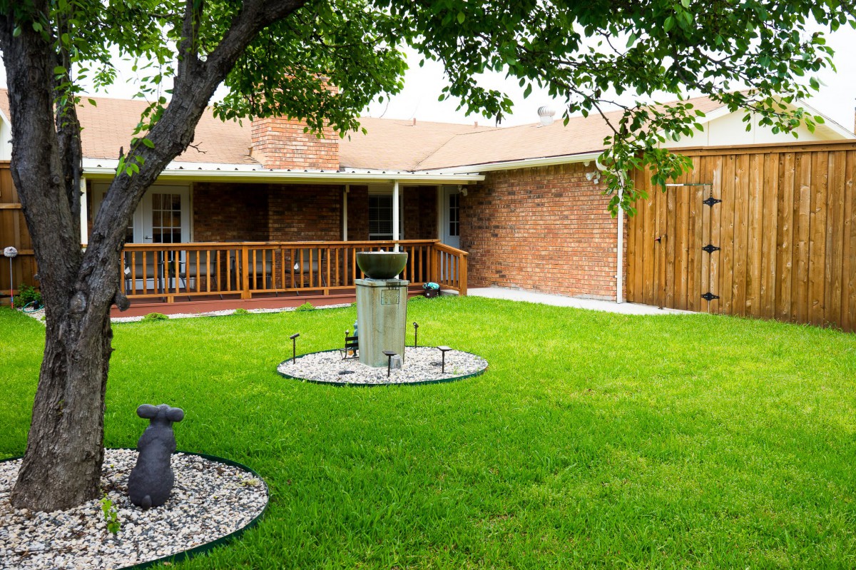 A well-maintained backyard with a lush green lawn, a tree, and a wooden deck attached to a brick house.