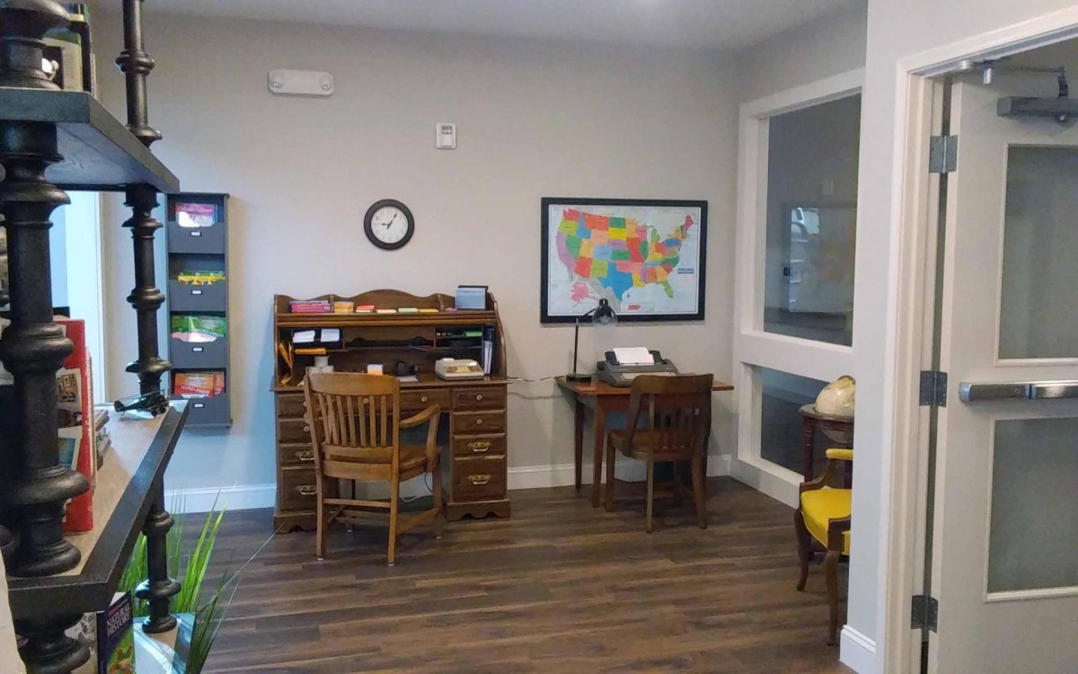 A small office space with wooden desks and chairs, featuring a map on the wall and a clock above.